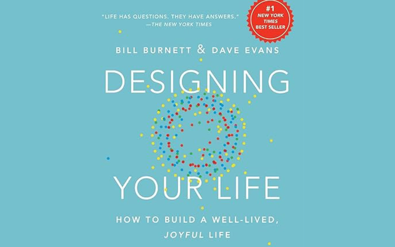 Hardcover book titled "Designing Your Life: How to Build a Well-Lived, Joyful Life" by Bill Burnett & Dave Evans. The blue cover features the words “DESIGNING YOUR LIFE” in large white letters, overlaid with concentric circles of yellow dots on the outside, blue dots for next ring, and red dots for the innermost ring.